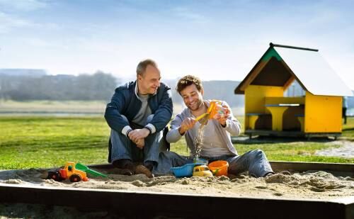 Sujet zur Kampagne Mehr Zeit für die Familie: Älterer Vater und erwachsener Sohn sitzen in der Sandkiste und spielen