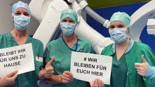 Spitalsmitarbeiterinnen mit Schutzmasken und Hinweistafeln mit der Aufschrift 'Wir bleiben für euch hier – bleibt ihr für uns zuhause'.