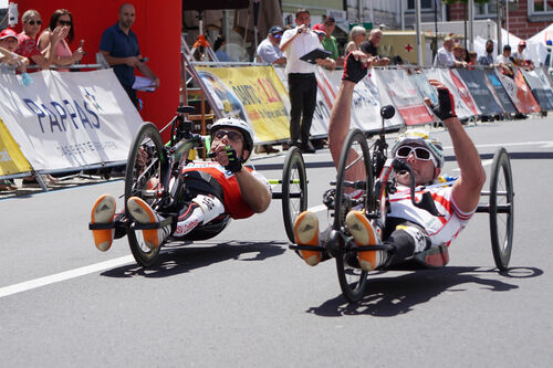 Zwei Handbike-Fahrer im Rennen nebeneinander, im Hintergrund Absperrung und Zuschauer