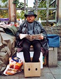 Wohnungsloser Mann sitzt auf einer Bank, liest Zeitung und ist umgeben von seinen Habseligkeiten 