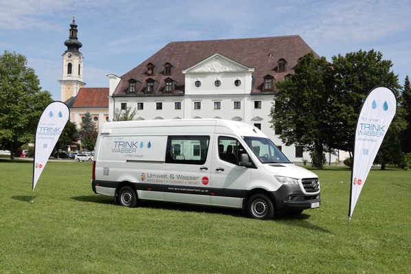 Ein weißer Bus mit der Aufschrift „Für unser Trinkwasser unterwegs“ steht gut sichtbar auf einer Wiese. Zwei Fahnen mit dem gleichen Motiv flankieren den Bus.