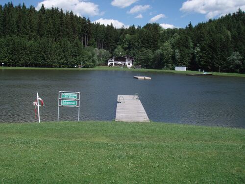 Blick auf den Badeseee Pramet mit langem Holzsteg mit zwei Leitern und einer schwimmenden Holzinsel