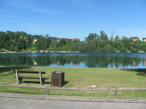 Badesee Regau in dem sich Bäume und Häuser spiegeln, im Vordergrund eine einsame Holzbank