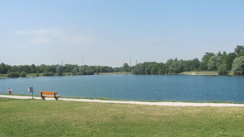 dunkelblauer Badesee  in Traun-Oedt, im Vodergrund eine leere Bank mit daneben stehendem Abfalleimer