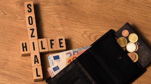 Neben Holzbuchstaben Sozialhilfe liegt eine Geldtasche, die mehrere Euroscheine und Euromünzen beinhaltet.