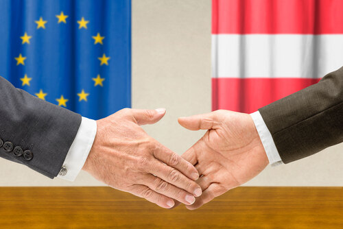 Zwei Männer schütteln sich die Hände, im Hintergrund sind die EU-Flagge und die Österreich-Flagge abgebildet