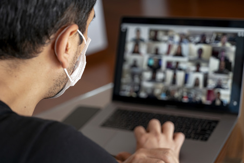 Mann sitzt vor einem Notebook bei einer Videokonferenz und trägt eine MNS-Maske 