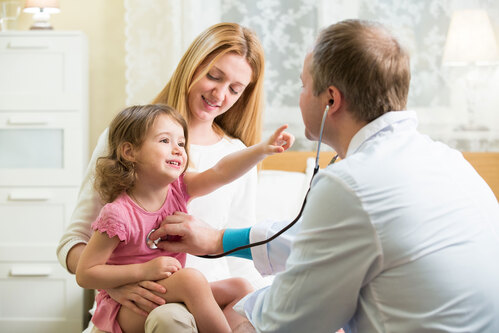 Ein Mädchen sitzt am Schoß seiner Mutter und wird von einem Arzt mit einem Stethoskop abgehört.