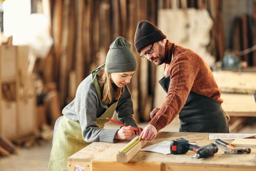 Eine Tischlerin und ein Tischler arbeiten an einem Holzdetail in einer Werkstatt.