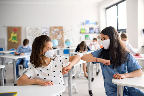 Schülerinnen sitzen mit Mundschutz in einer Klasse nebeneinander und begrüßen sich mit ihren Ellbogen