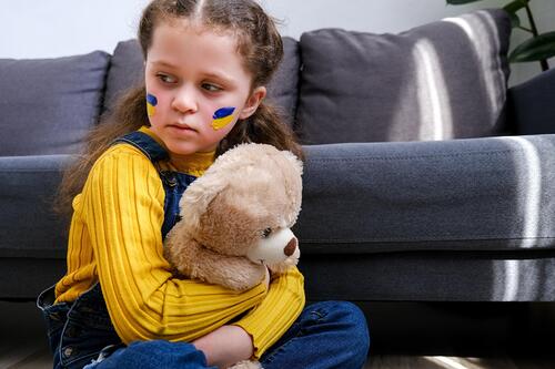 Ukrainisches Flüchtlingskind sitzt mit einem Teddybär vor einem Sofa