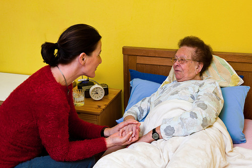 Junge Frau sitzt bei einer älteren, pflegebedürftigen Frau am Bett