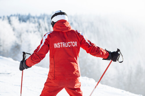 Skilehrer steht auf einer Skipiste und trägt eine rote Skijacke mit der Aufschrift 'Instructor'