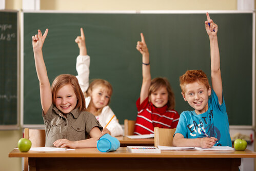 Vier Volksschüler sitzen in der Klasse auf der Schulbank und zeigen auf 