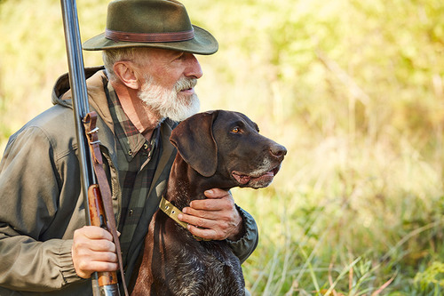 Jäger sitzt mit seinem Jagdhund in einer Wiese.